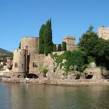 Château médiéval en pierre situé au bord d'une étendue d'eau, entouré d'arbres et de végétation, avec une plage et des personnes en arrière-plan