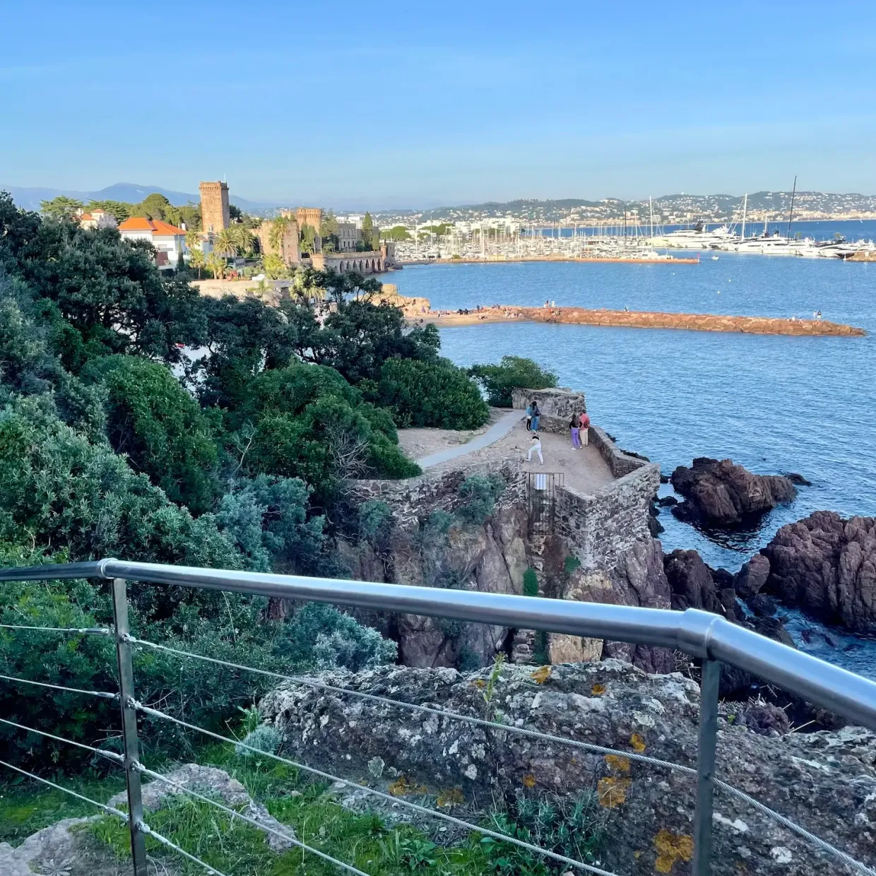 Vue sur un port de plaisance avec des bateaux, une forteresse médiévale et des promeneurs sur un sentier côtier