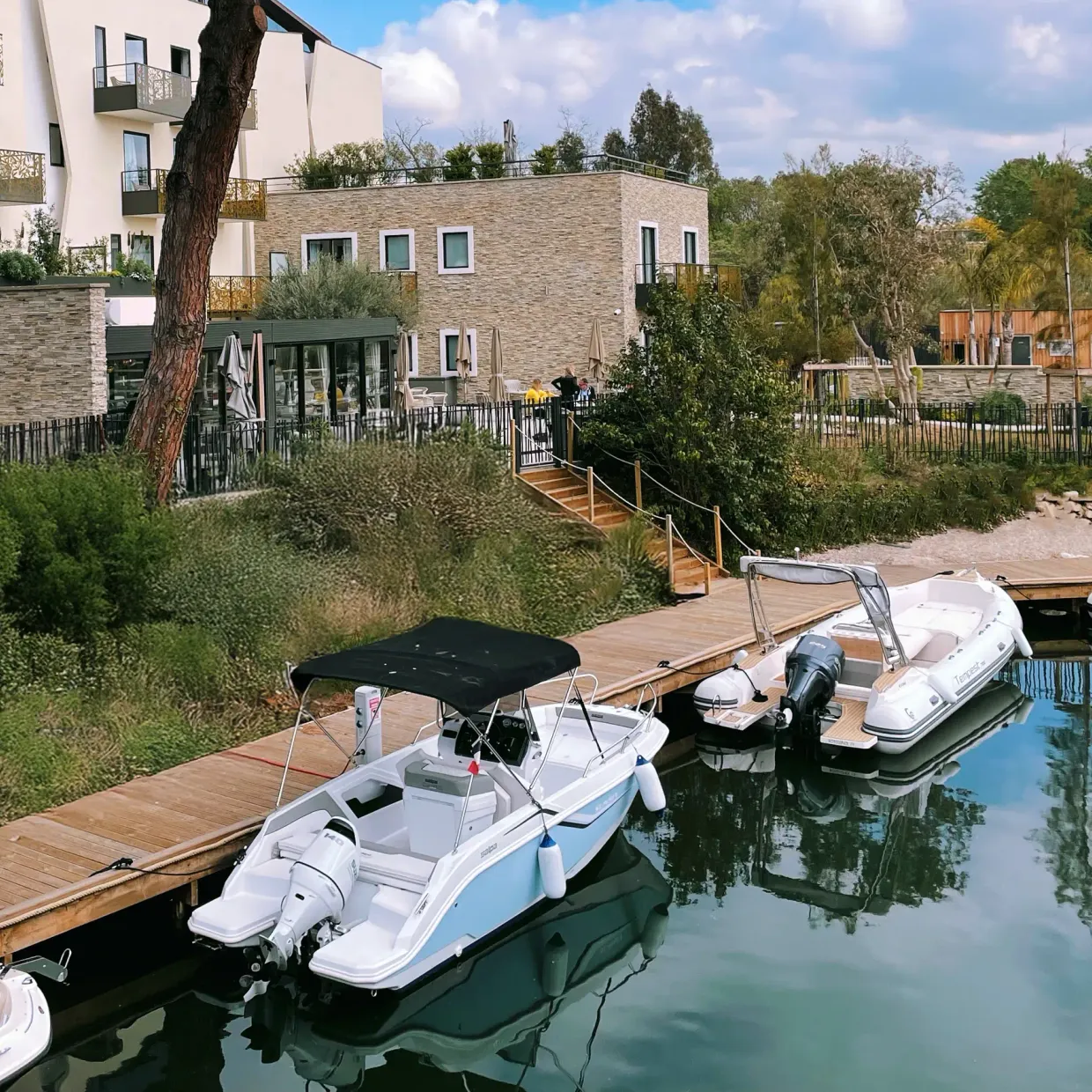 Quais en bois avec plusieurs petits bateaux à moteur amarrés devant des bâtiments résidentiels modernes entourés de végétation