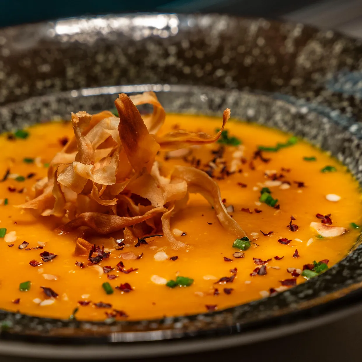 Un bol contenant une soupe orange garnie de chips et d'herbes fraîches, posé sur une surface sombre