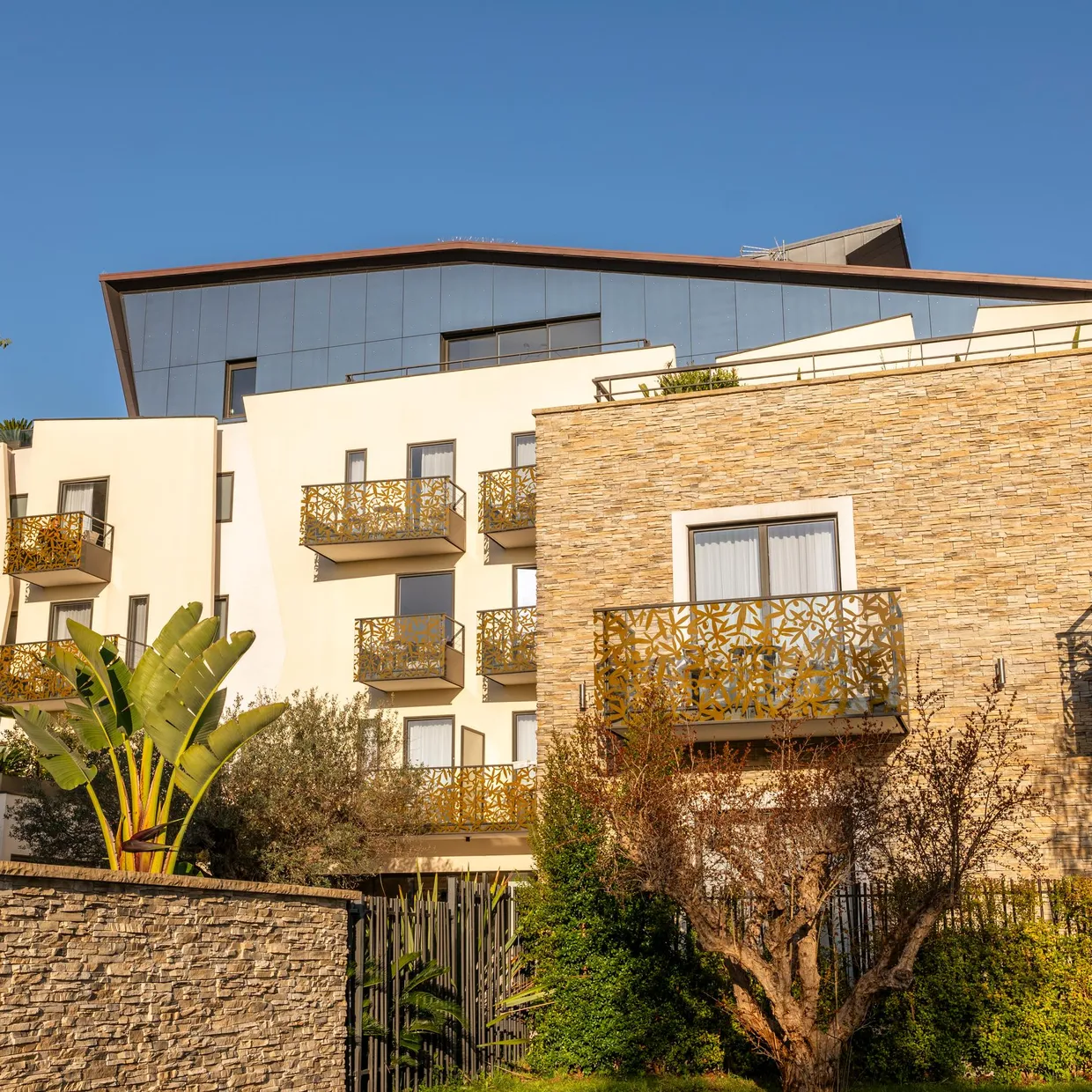 Immeuble moderne avec balcons ornés de motifs en métal et façade en pierre, entouré d'une végétation variée