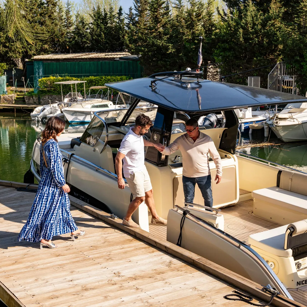 Trois personnes montent à bord d'un bateau amarré dans un port entouré d'autres embarcations et de végétation