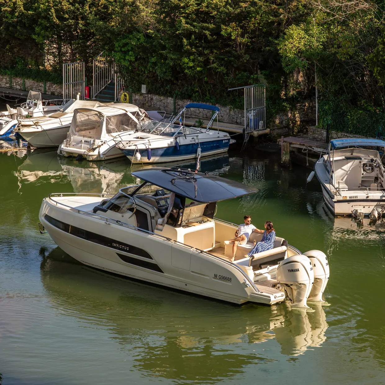 Un bateau de plaisance blanc avec deux personnes à bord est amarré dans un petit port entouré d'autres embarcations et de végétation