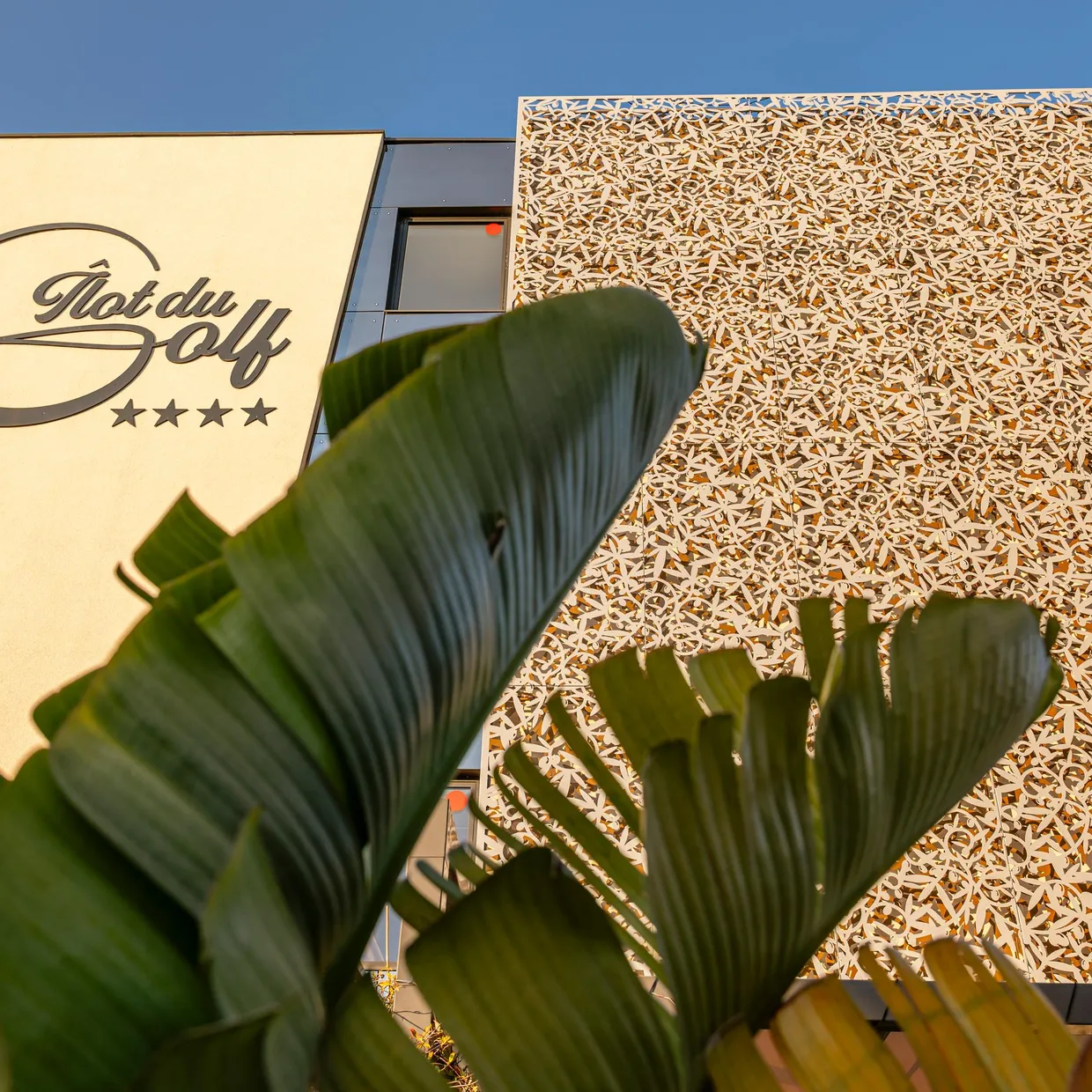 Façade d'un bâtiment avec l'enseigne "Hôtel du Golf" et quatre étoiles, partiellement masquée par des feuilles de plante