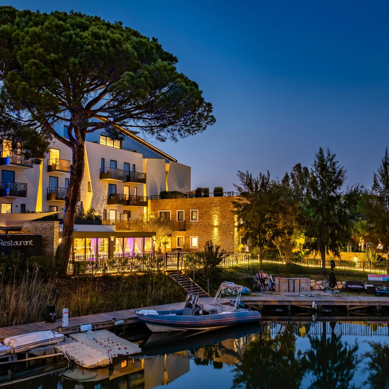 Un bâtiment moderne éclairé avec un restaurant au rez-de-chaussée, entouré d'arbres et situé près d'un plan d'eau avec des bateaux amarrés