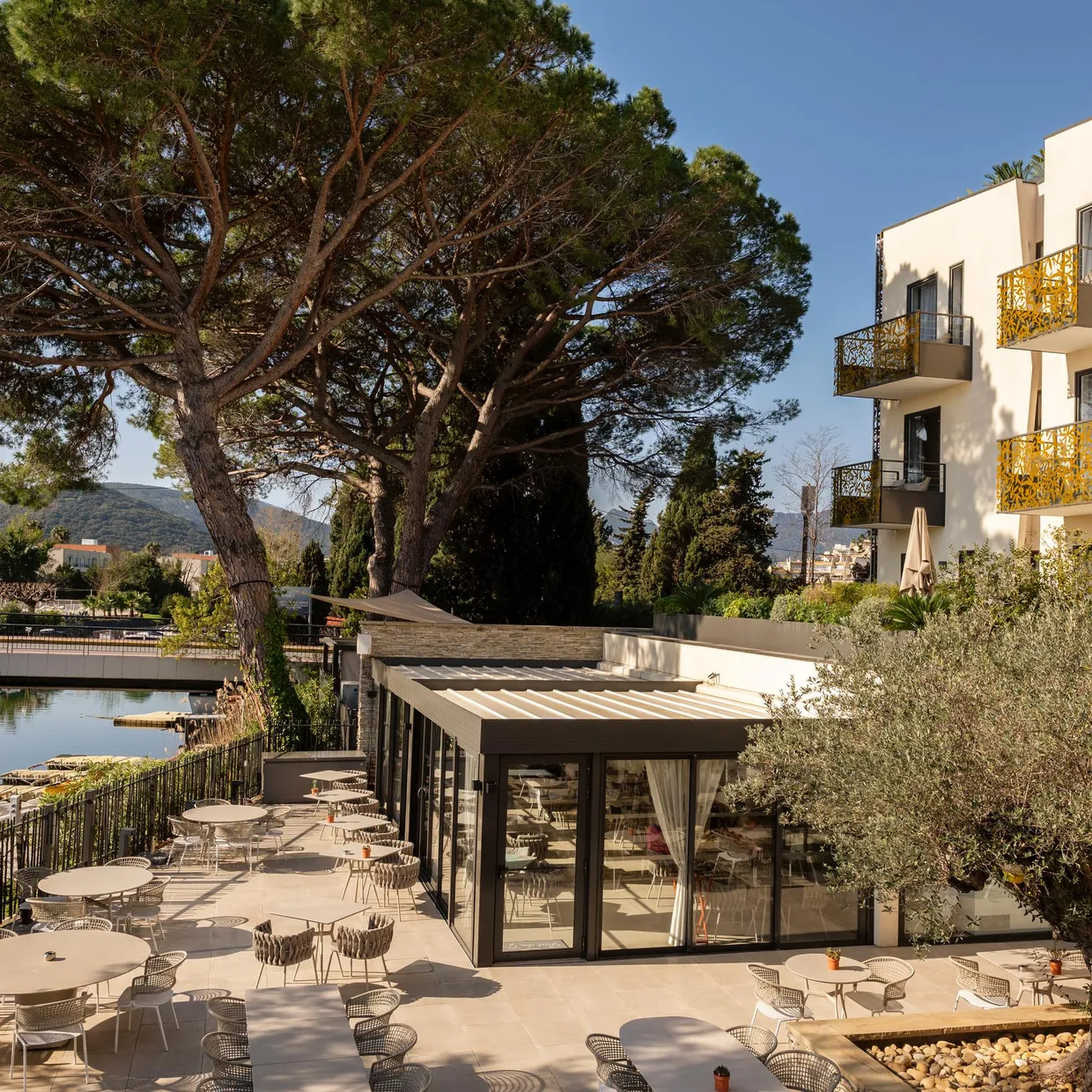 Terrasse de restaurant avec tables et chaises près d'un plan d'eau, entourée d'arbres et de bâtiments modernes