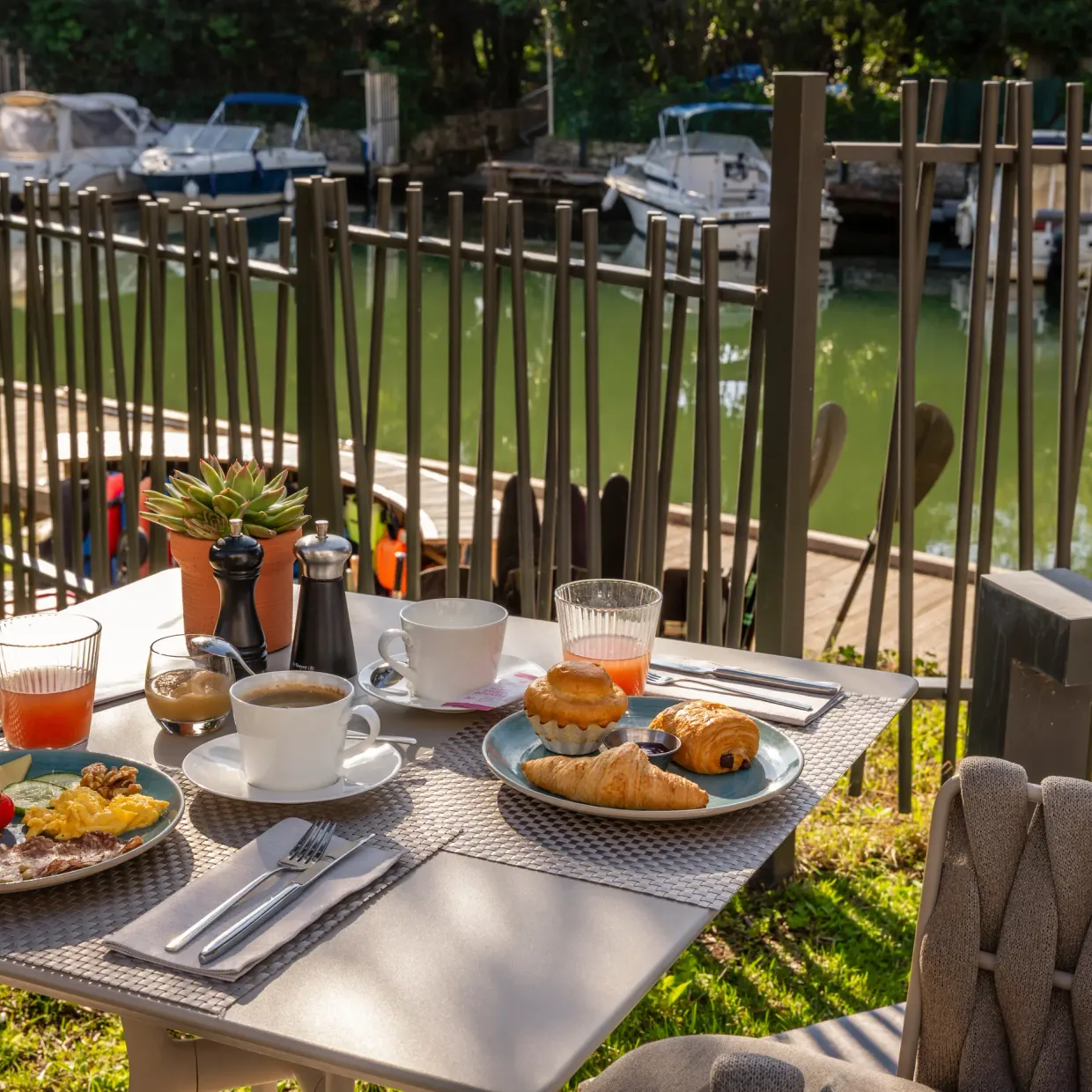 Table en extérieur avec petit-déjeuner composé de croissants, pain, confiture, fruits et boissons, vue sur un canal avec des bateaux