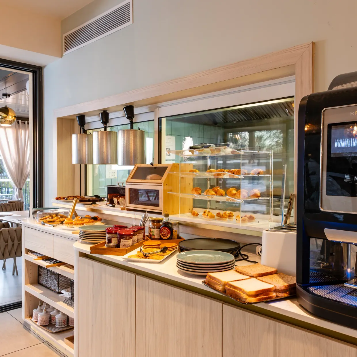 Un comptoir de petit-déjeuner avec des viennoiseries, des toasts, des confitures et une machine à café, dans une salle lumineuse avec des tables et des chaises à proximité