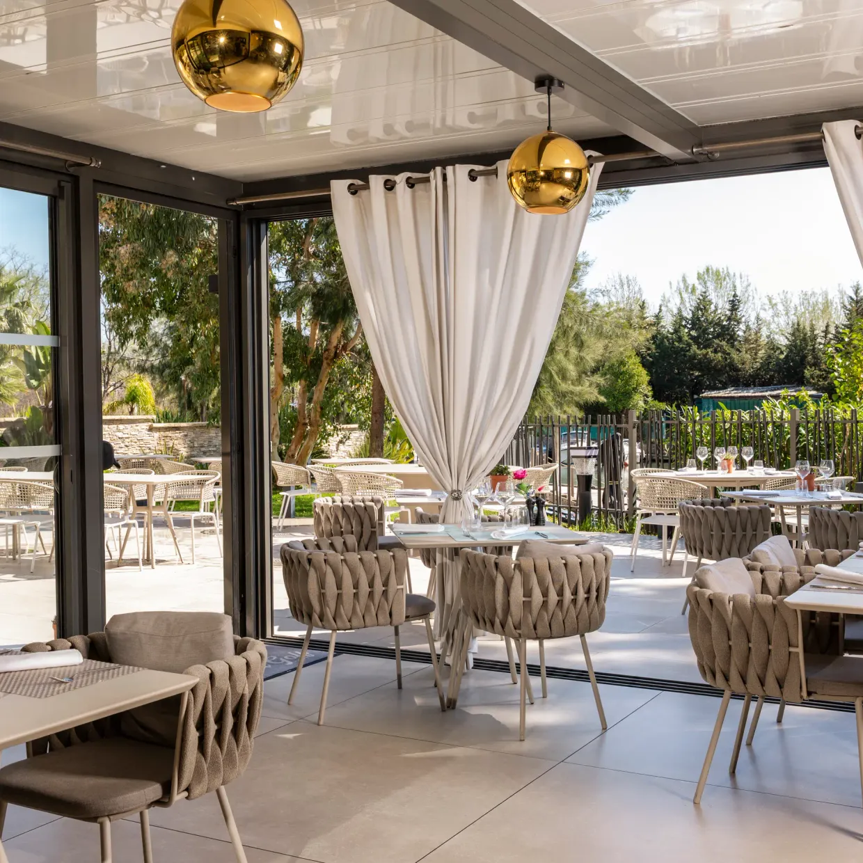 Salle de restaurant avec tables et chaises disposées, grandes baies vitrées ouvertes sur une terrasse extérieure, décorée de plantes et de luminaires sphériques