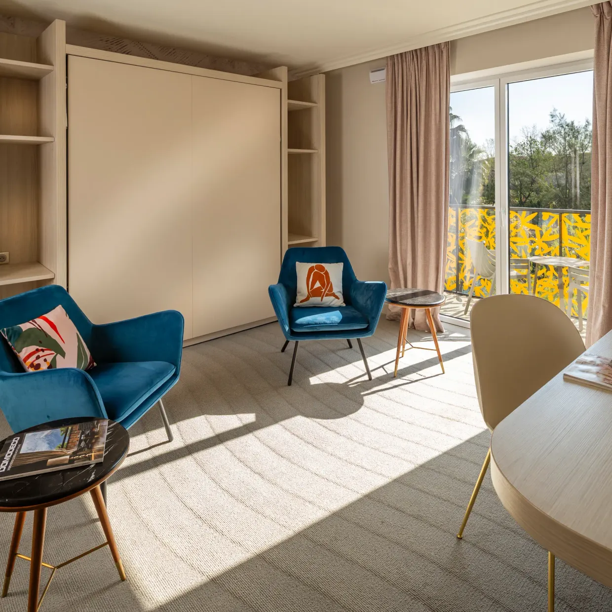 Pièce avec deux fauteuils bleus, une table ronde, un bureau, des étagères et une porte-fenêtre donnant sur un balcon avec une vue sur des arbres
