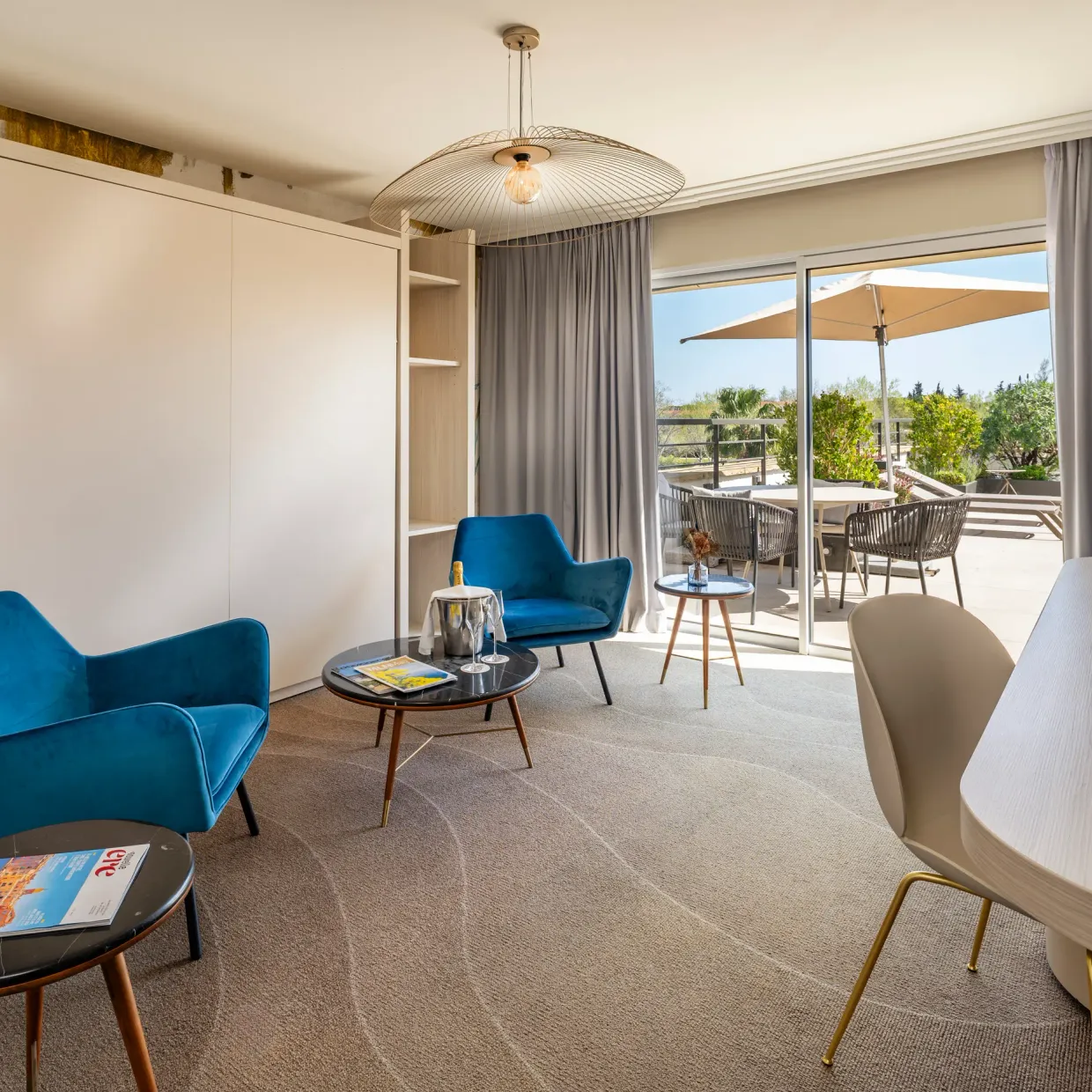 Salon moderne avec deux fauteuils bleus, une table basse, un bureau et une vue sur une terrasse extérieure avec des chaises et des parasols