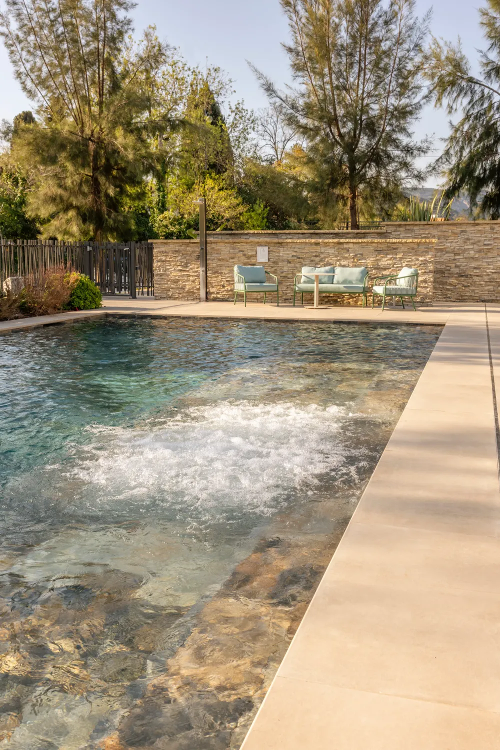 Piscine avec un coin de détente composé de chaises longues et d'une table basse, entourée d'arbres et d'une clôture en pierre