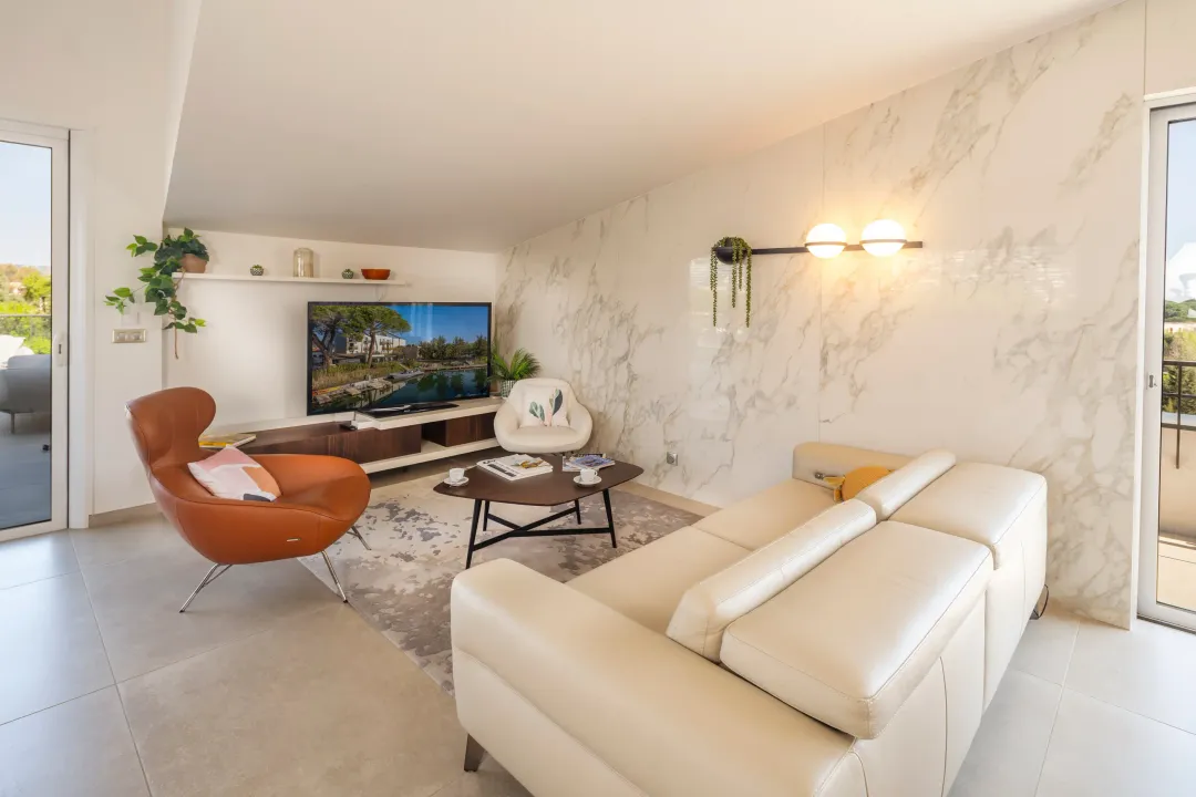 Salon moderne avec canapé beige, fauteuil en cuir marron, table basse et télévision affichant un paysage