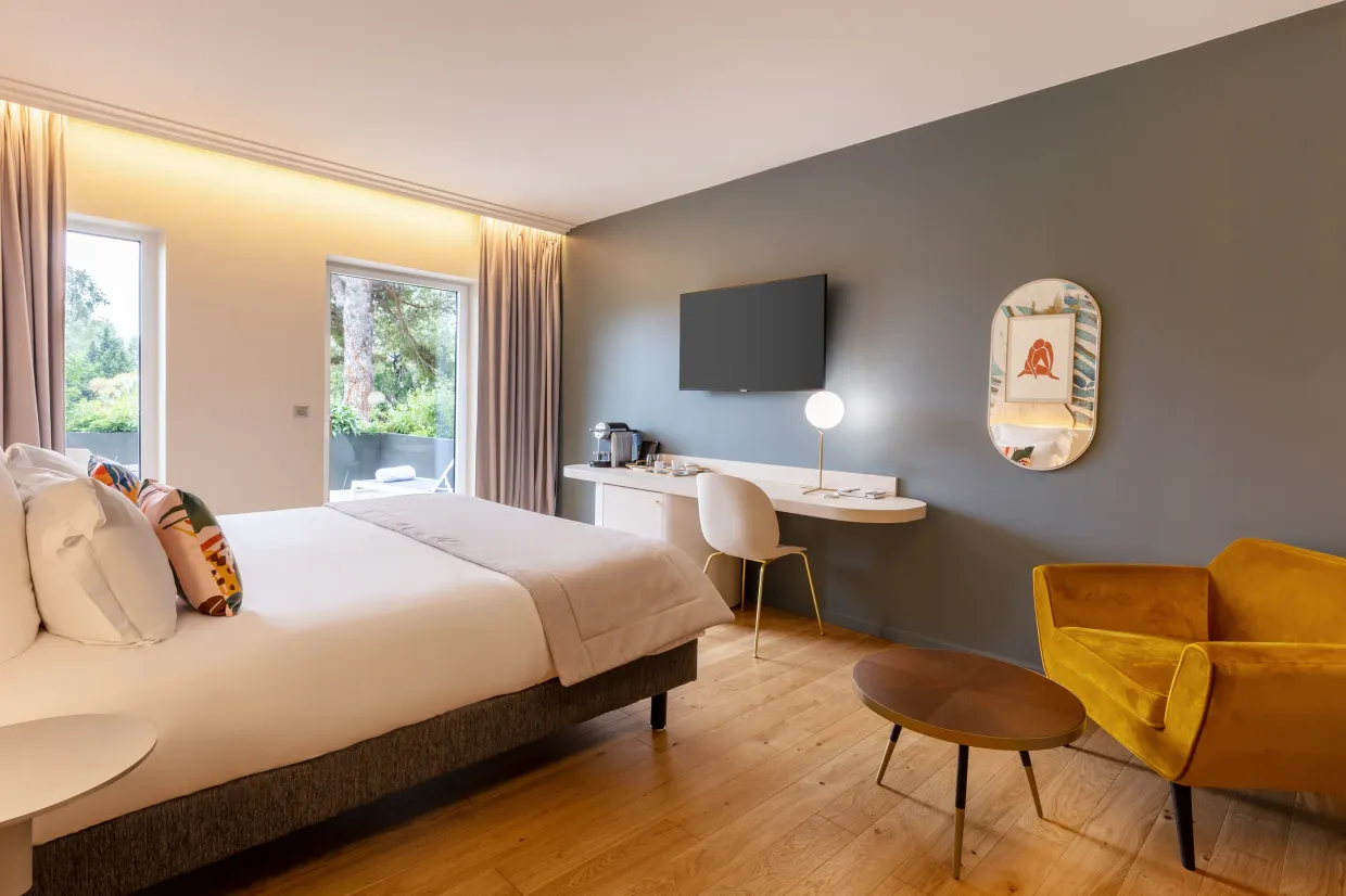 Chambre moderne avec un lit, une télévision, un bureau, un fauteuil jaune et une table basse, éclairée par la lumière naturelle des fenêtres
