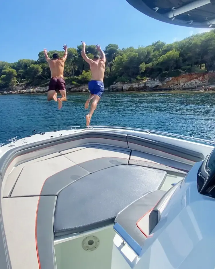 Deux hommes sautent depuis l'arrière d'un bateau dans une mer bleue entourée de végétation et de rochers