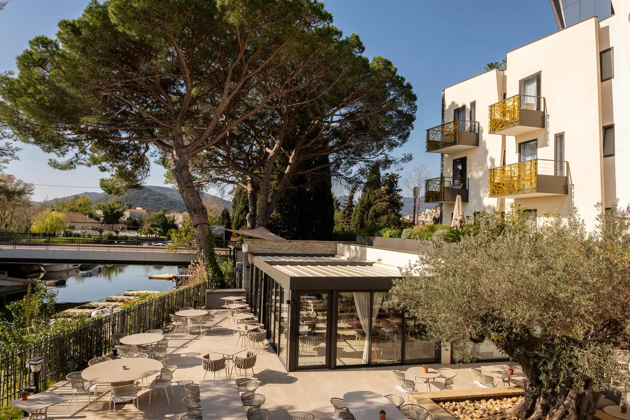Terrasse de restaurant avec tables et chaises près d'un plan d'eau, entourée d'arbres et de bâtiments modernes