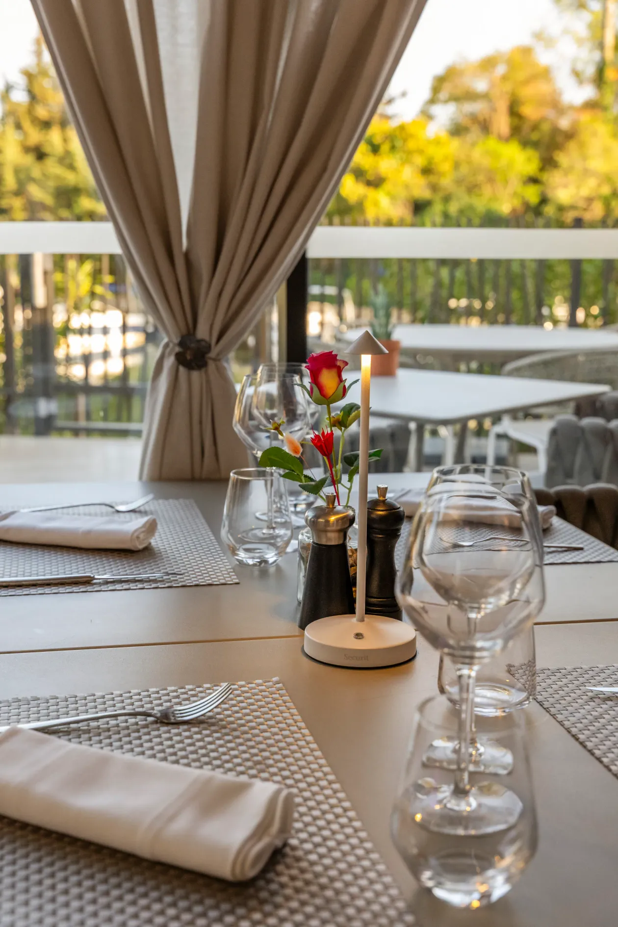 Table dressée avec des verres à vin, des serviettes, des fleurs et des moulins à poivre, vue depuis un restaurant avec des arbres en arrière-plan