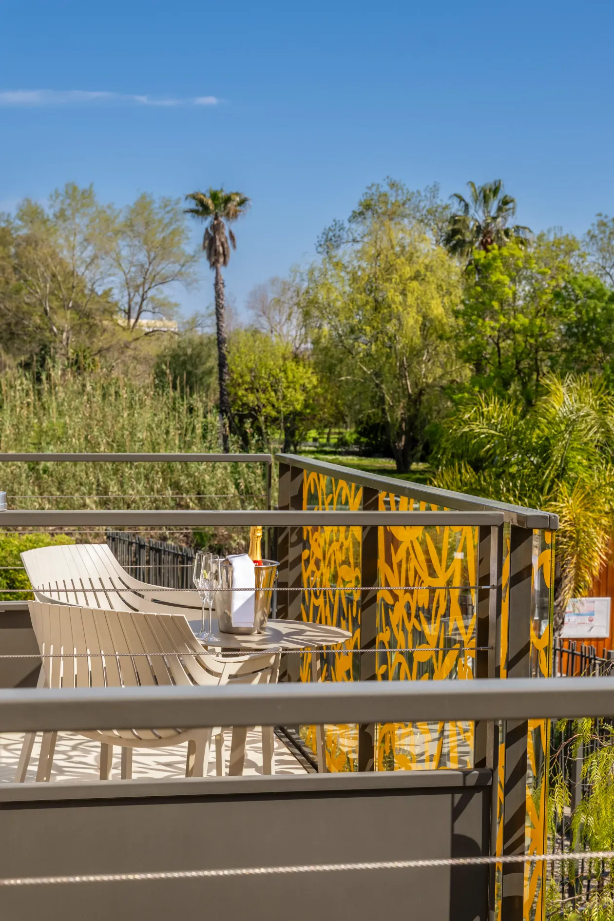 Deux chaises longues et une table avec des verres et un seau à champagne sur un balcon avec une rambarde métallique et un décor jaune, entourées d'arbres et de végétation