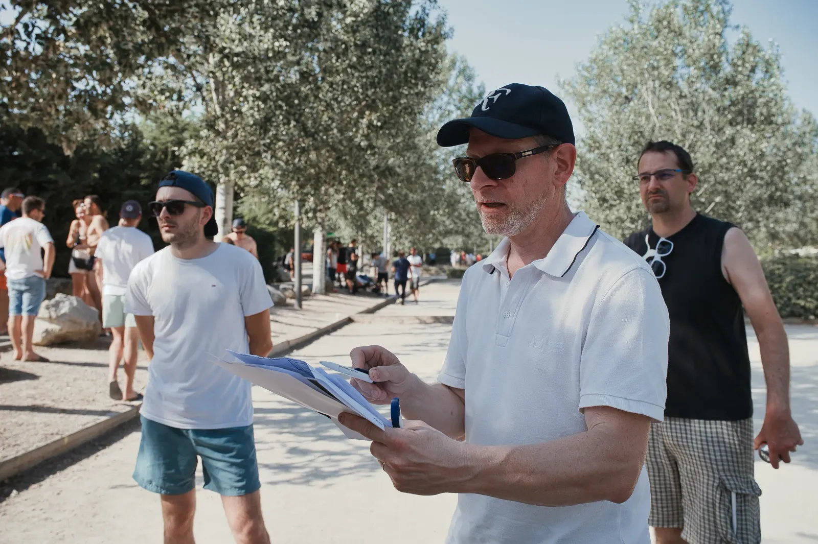 Un homme en chemise blanche et casquette lit des documents en plein air, entouré de personnes en tenue estivale