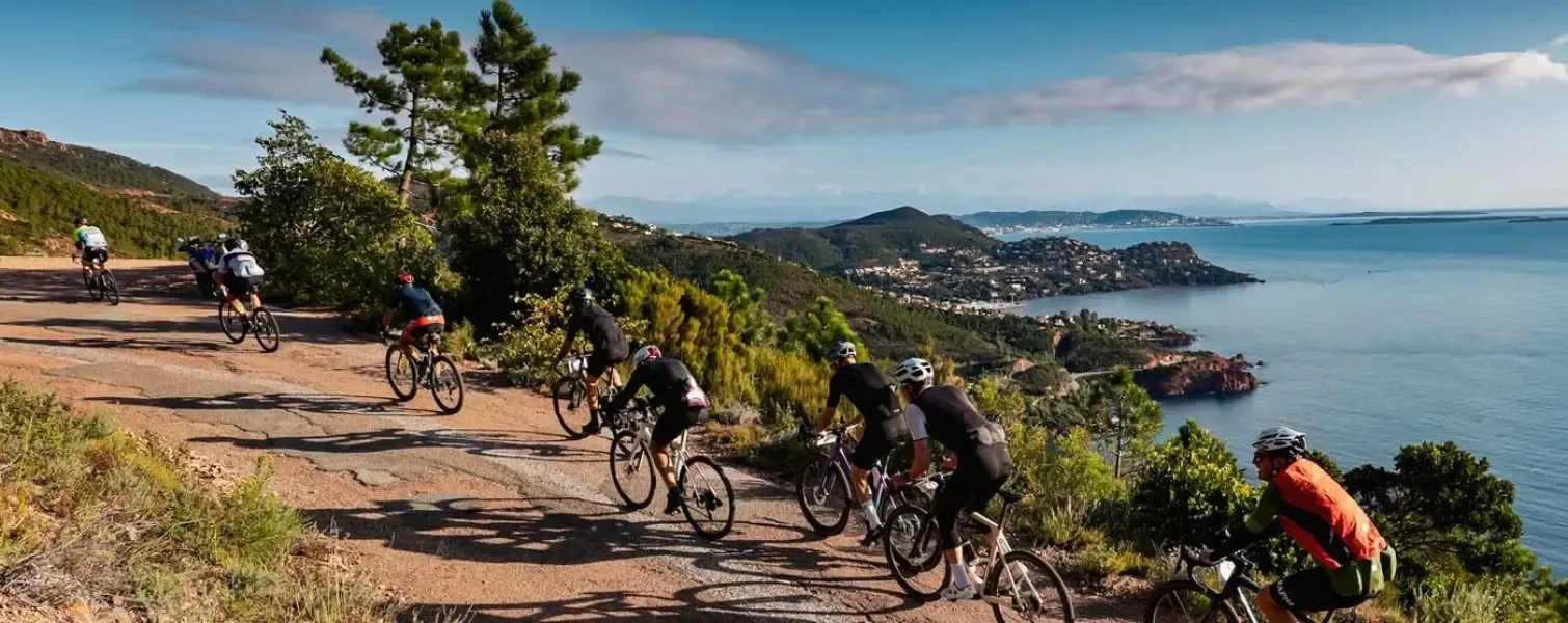 Un groupe de cyclistes pédale sur un chemin de terre en bord de mer avec une vue sur une ville côtière et des collines