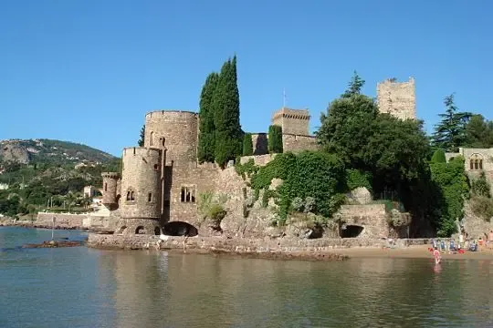 Château médiéval en pierre situé au bord d'une étendue d'eau, entouré d'arbres et de végétation, avec une plage et des personnes en arrière-plan