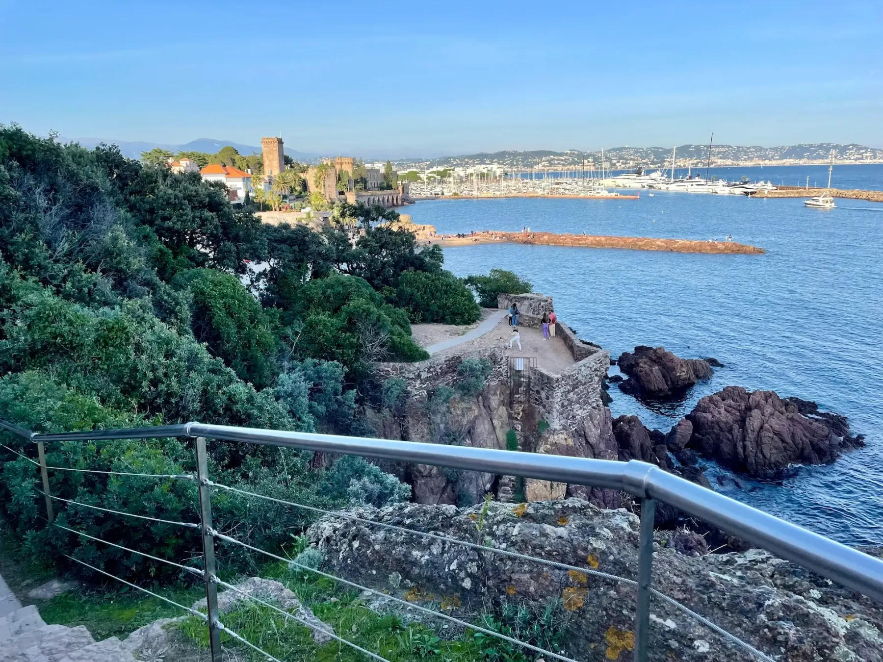 Vue sur un port de plaisance avec des bateaux, une forteresse médiévale et des promeneurs sur un sentier côtier