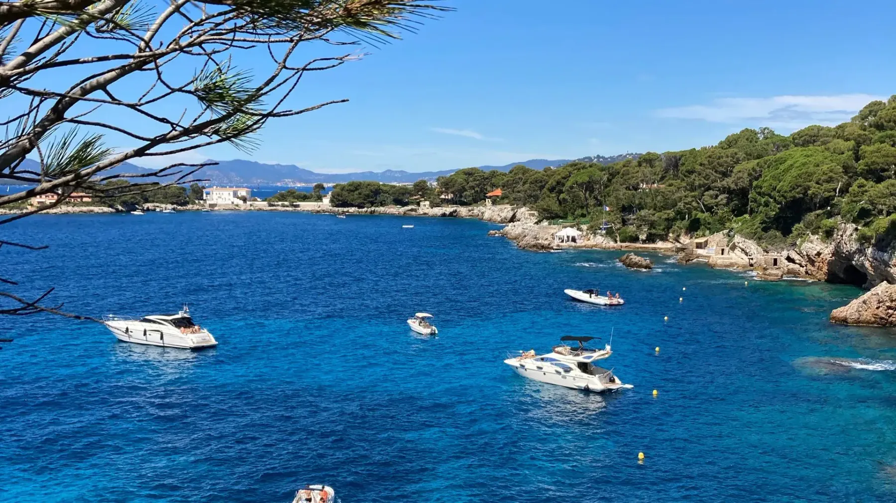 Plusieurs bateaux naviguent dans une baie aux eaux turquoise bordée de végétation et de rochers, avec des montagnes en arrière-plan
