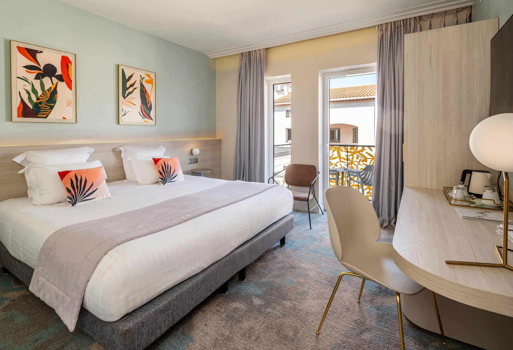 Chambre d'hôtel avec un lit double, une table de chevet, une chaise et une porte-fenêtre donnant sur un balcon avec une vue sur des toits