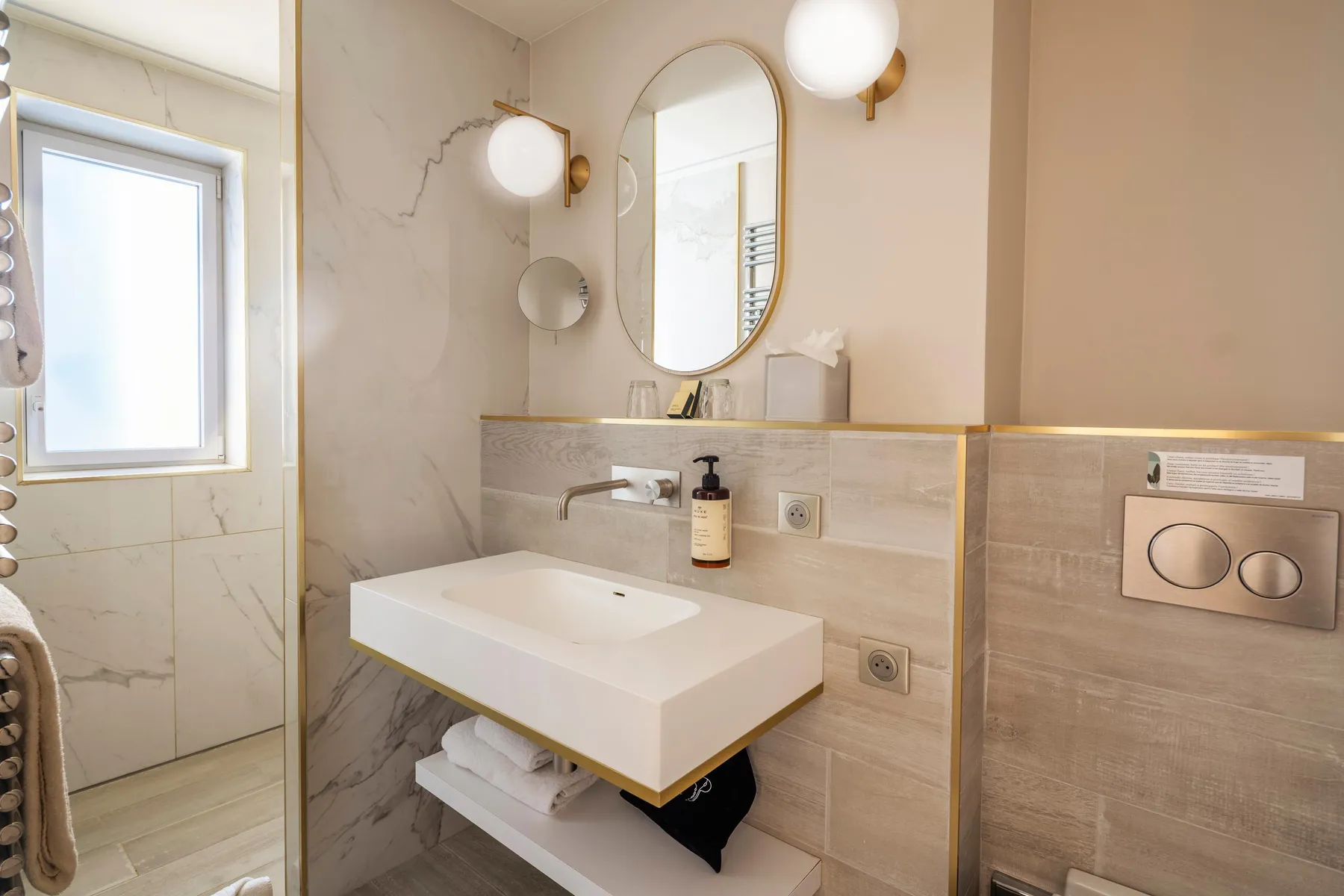 Un lavabo blanc avec robinetterie, des miroirs ovales et ronds, des lumières sphériques, une fenêtre et des étagères dans une salle de bain aux murs carrelés de beige et de marbre