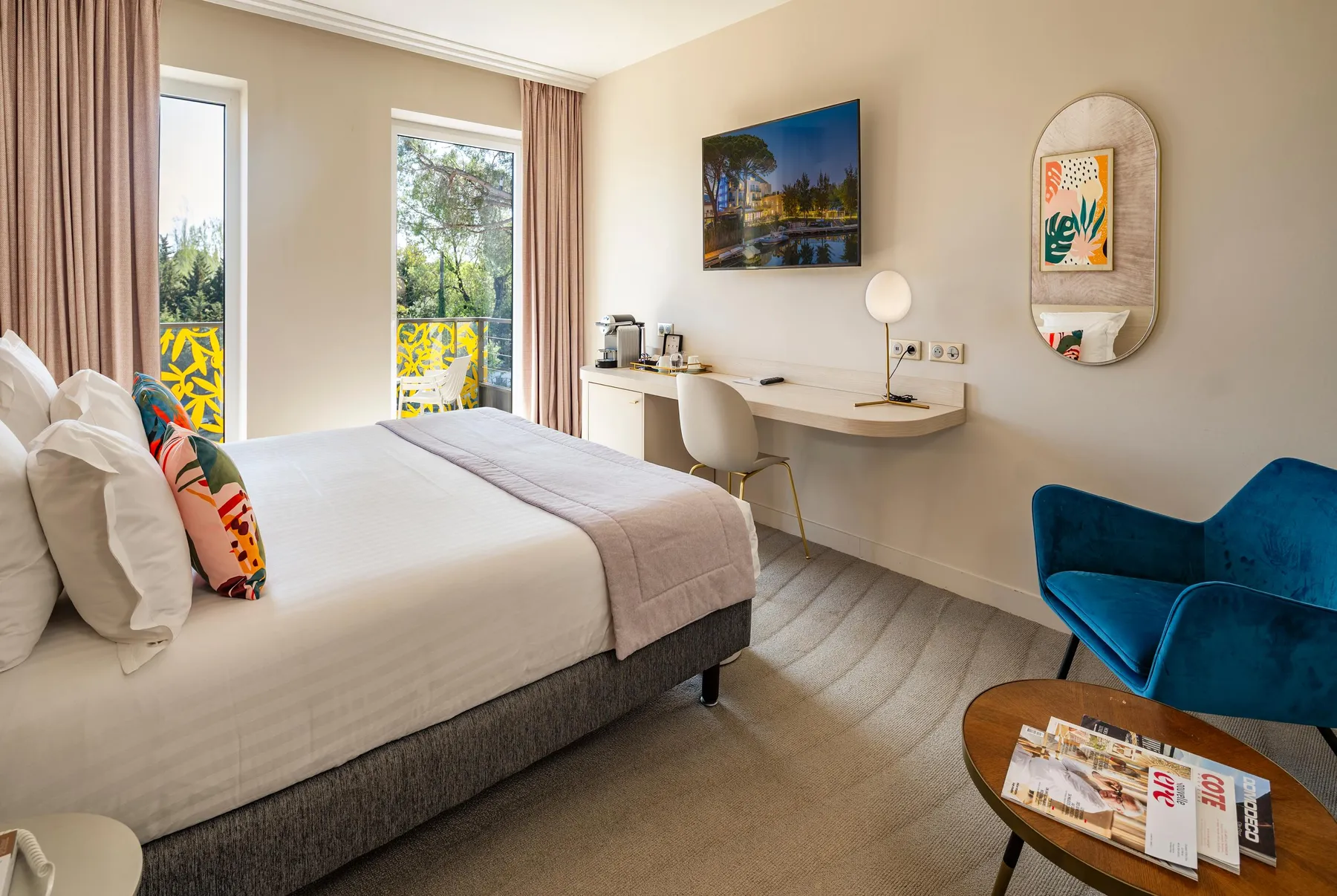 Chambre d'hôtel avec un lit, une table de travail, un fauteuil bleu, une télévision et une vue sur un balcon avec des motifs colorés