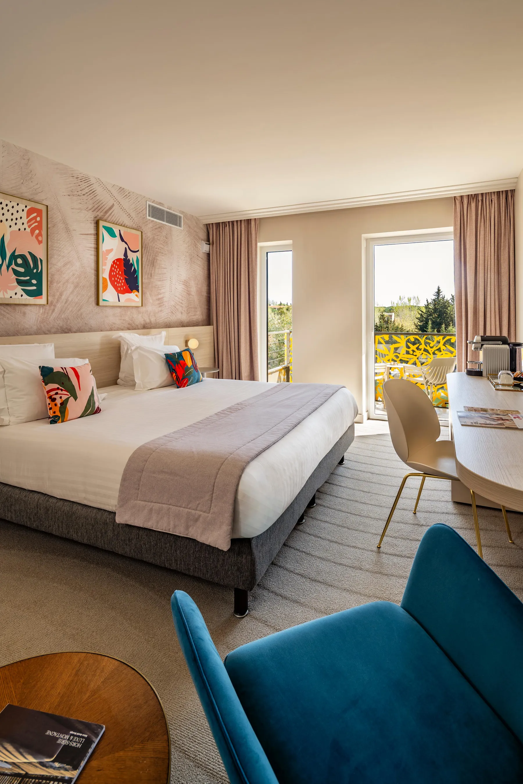Chambre d'hôtel avec un lit, des coussins colorés, deux fauteuils, une table avec une chaise et une porte-fenêtre donnant sur un balcon