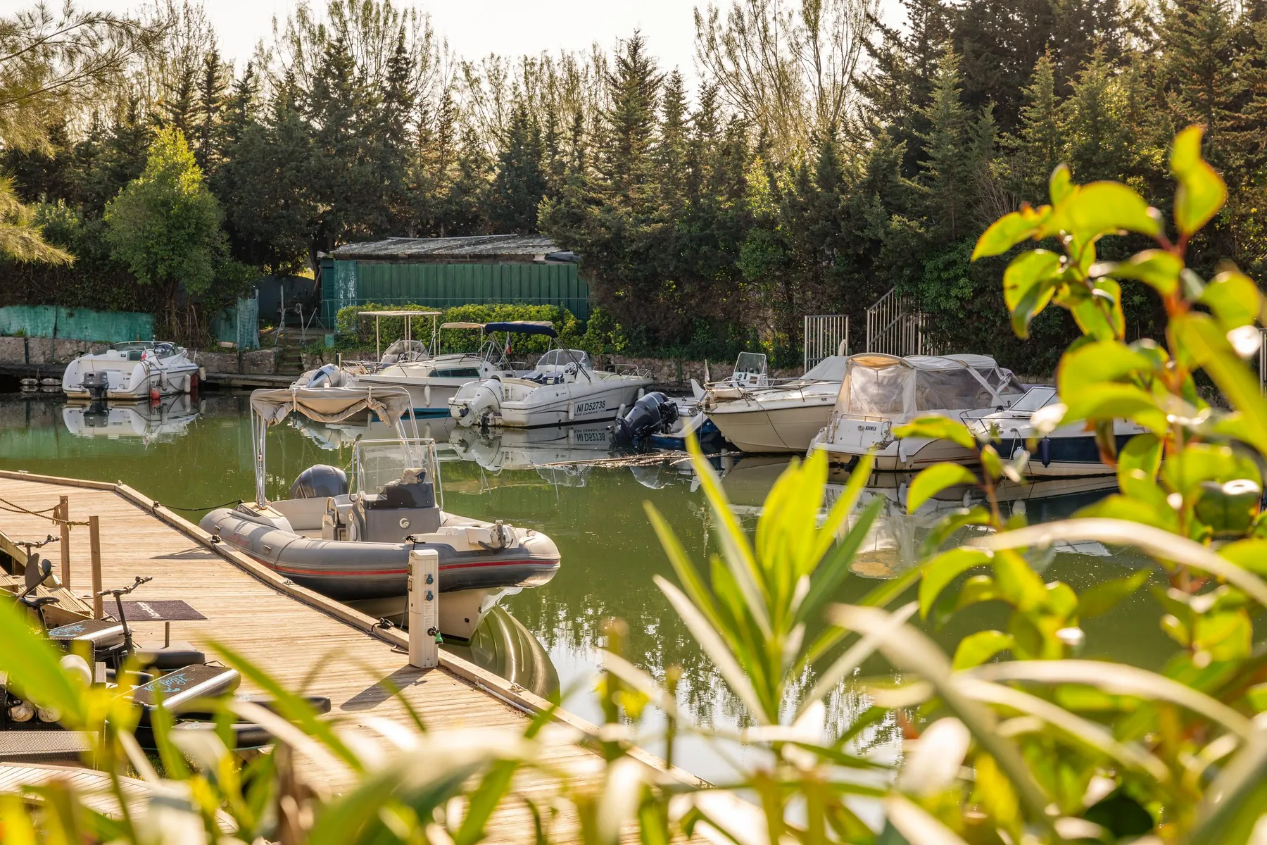 Plusieurs bateaux amarrés dans un canal entouré d'arbres et de végétation, avec un ponton en pierre au premier plan