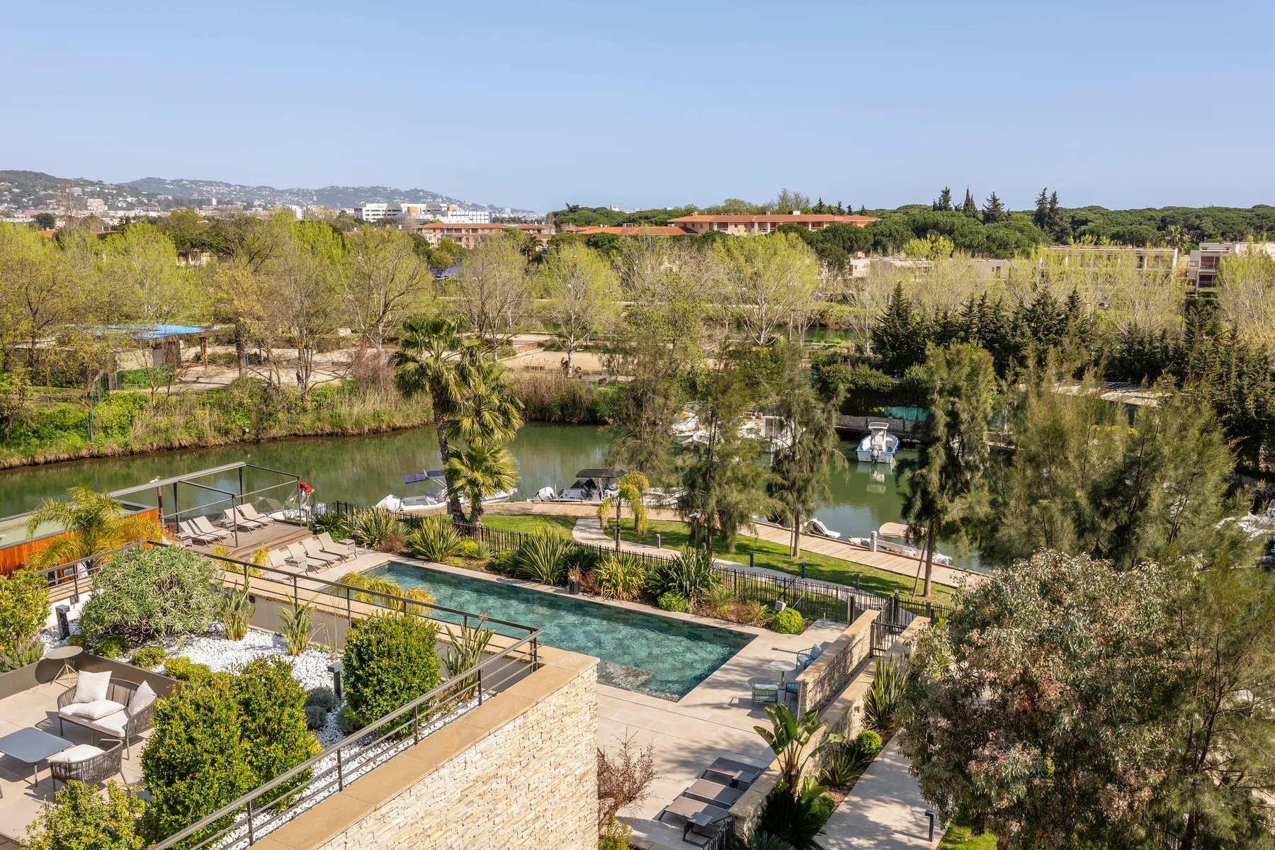 Vue aérienne d'un jardin avec piscine, bordé d'arbres et d'un plan d'eau où sont amarrés des bateaux