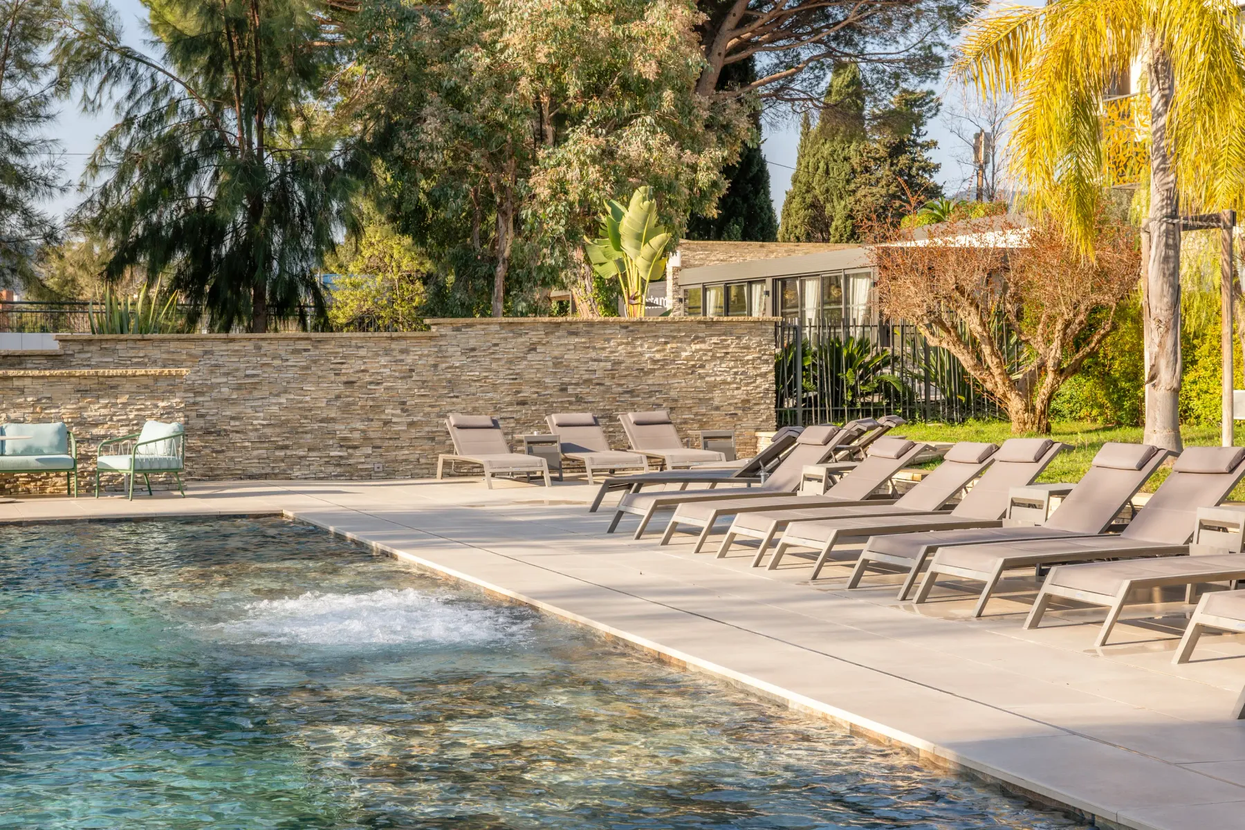 Une piscine avec des transats alignés et un mur de pierre en arrière-plan, entourée d'arbres et de végétation