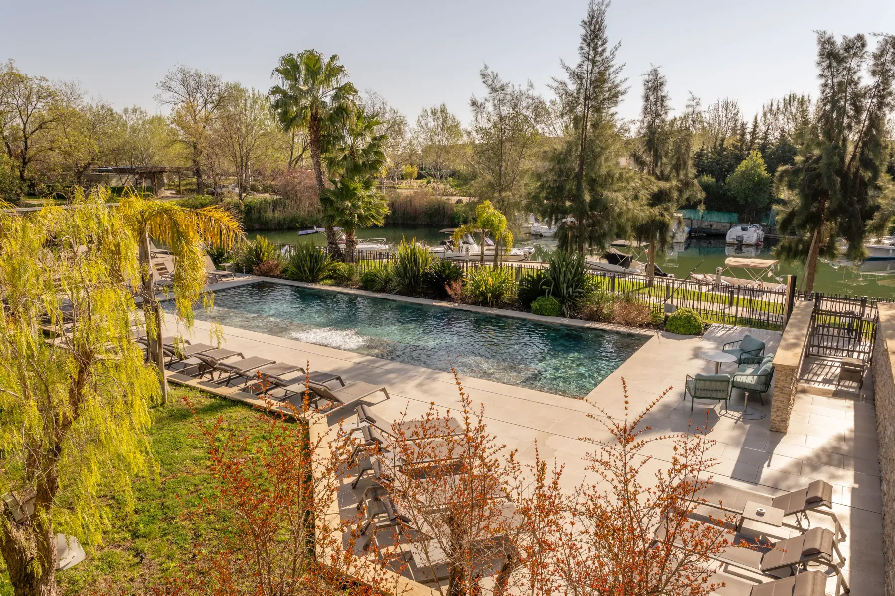 Piscine entourée de chaises longues et de tables, avec un plan d'eau et des bateaux en arrière-plan, dans un cadre arboré