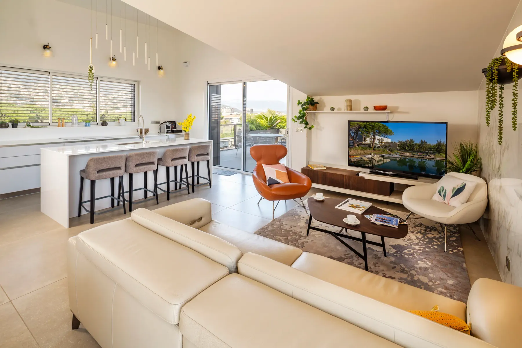 Salon moderne avec canapé blanc, fauteuils, table basse et télévision, adjacent à une cuisine avec îlot central et tabourets