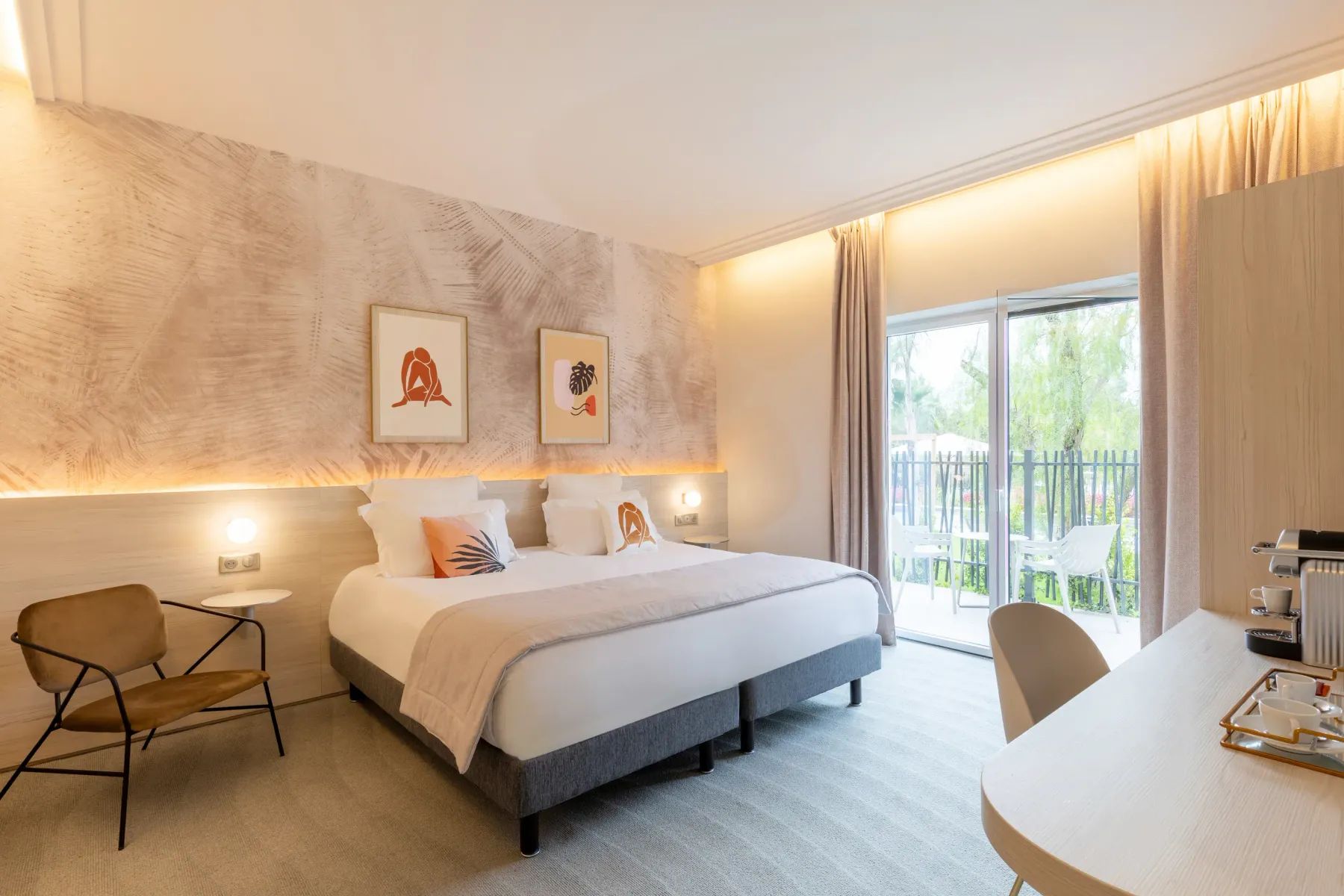 Chambre d'hôtel moderne avec un lit double, une table avec deux chaises, un fauteuil, une porte-fenêtre donnant sur un balcon et des œuvres d'art sur le mur
