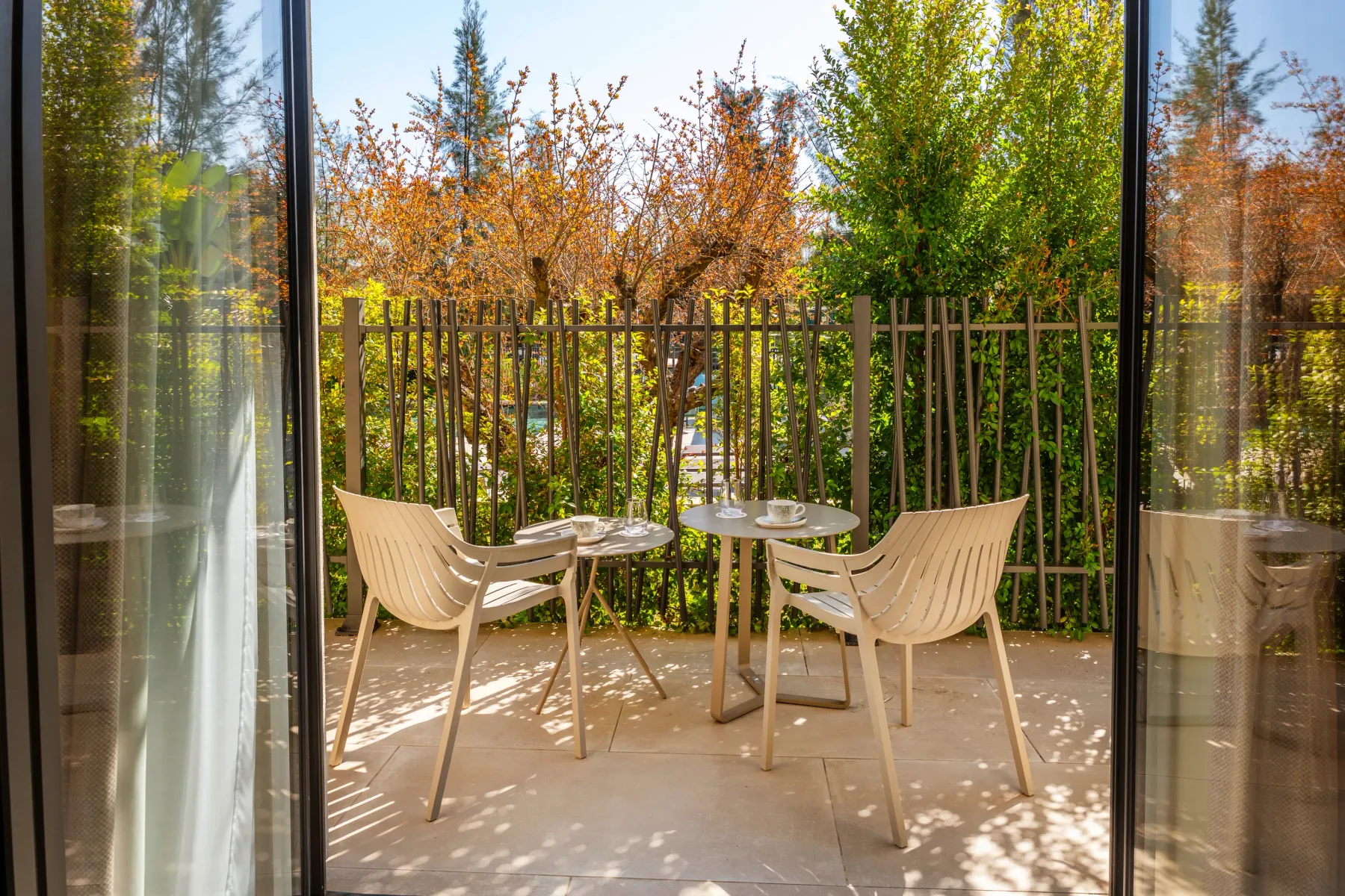 Deux chaises et une table rondes sur une terrasse en pierre, entourées d'une végétation dense et d'une clôture métallique, vues à travers une porte vitrée