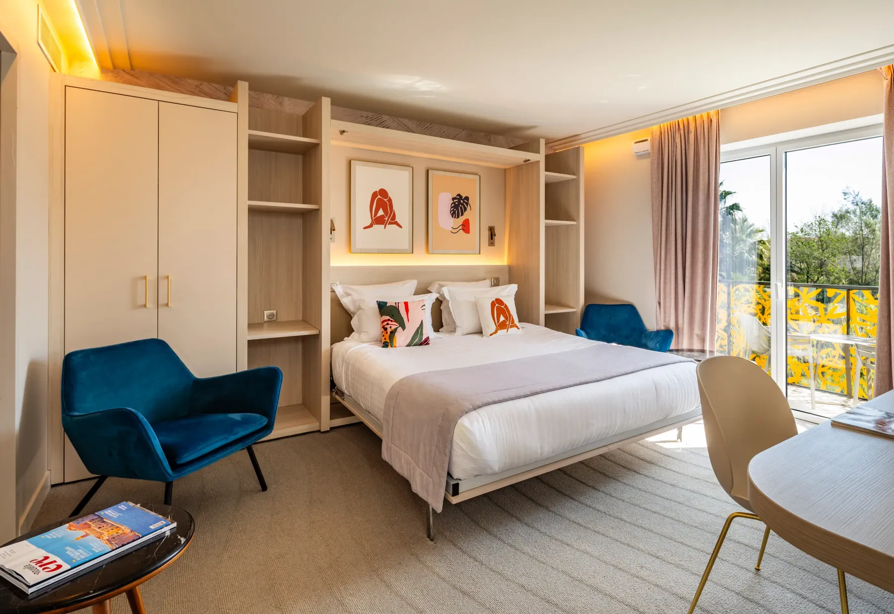Chambre d'hôtel avec un lit double, deux fauteuils, une table ronde et une vue sur un balcon avec des plantes