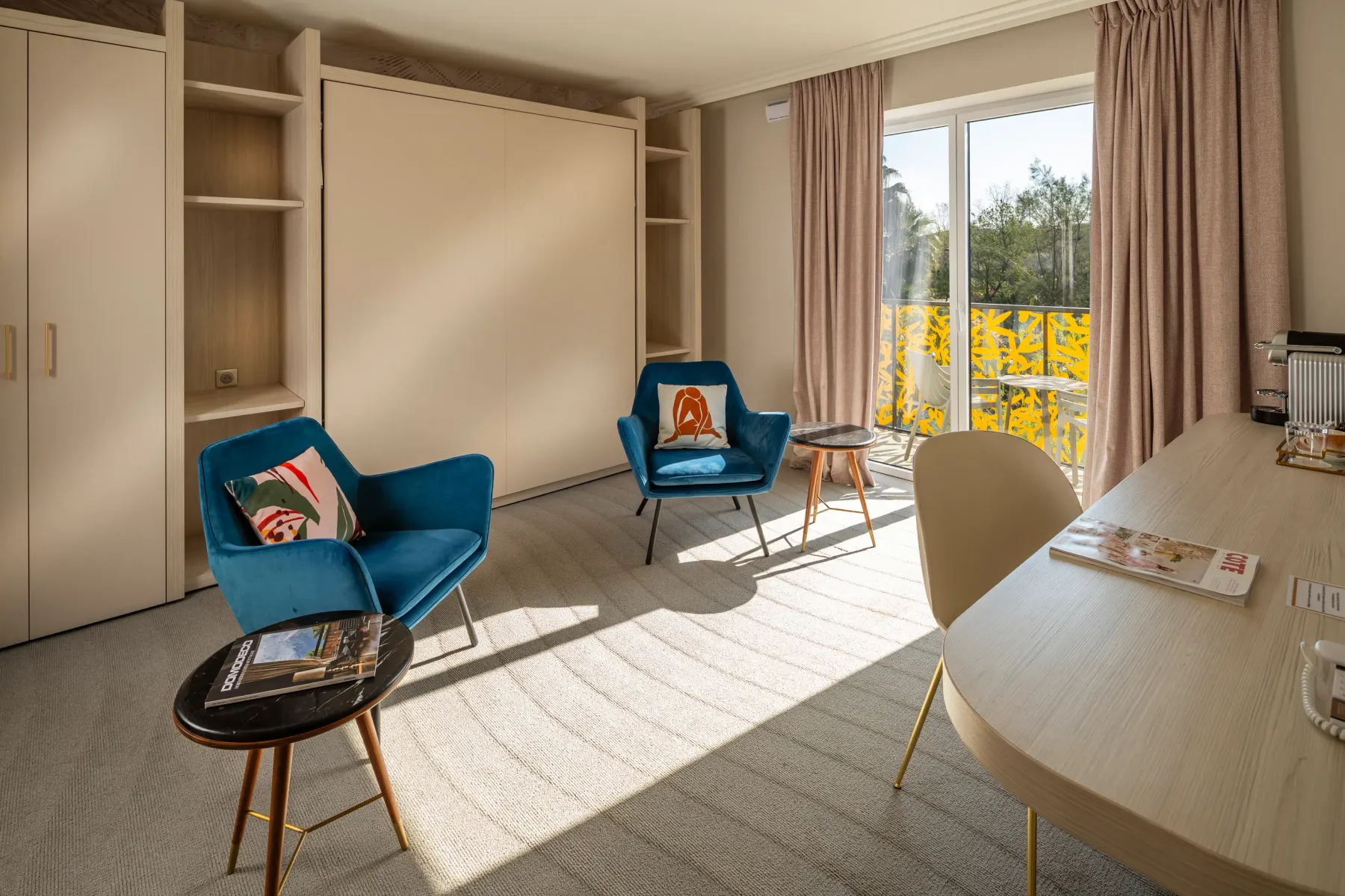 Pièce avec deux fauteuils bleus, une table ronde, un bureau, des étagères et une porte-fenêtre donnant sur un balcon avec une vue sur des arbres