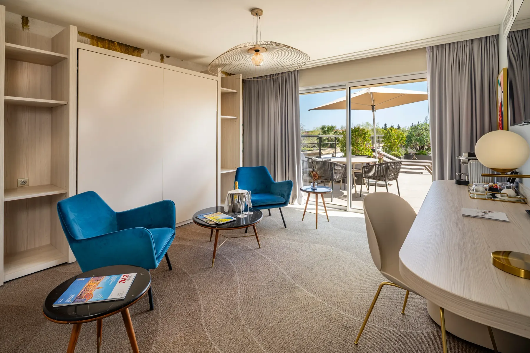 Salon moderne avec deux fauteuils bleus, une table basse, un bureau et une vue sur une terrasse extérieure avec des chaises et des parasols