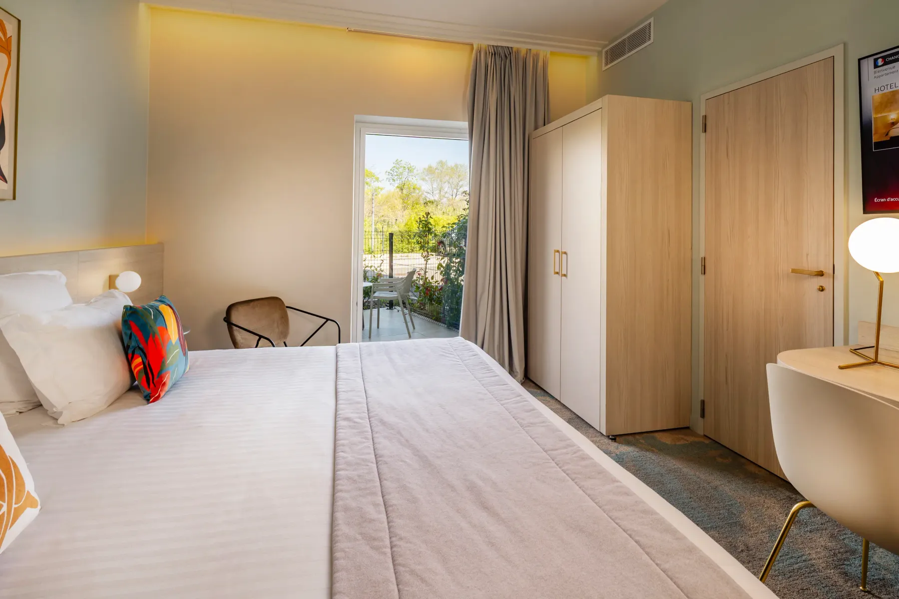 Chambre d'hôtel avec un lit double, une porte-fenêtre donnant sur un balcon, des armoires et une table de chevet avec une lampe