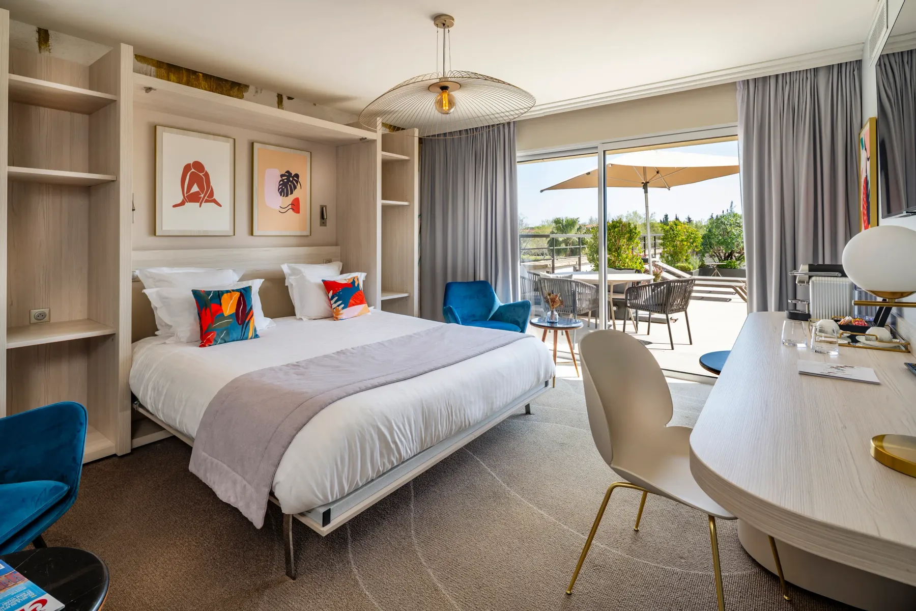 Chambre d'hôtel moderne avec un lit double, une table de travail, un fauteuil bleu et une vue sur une terrasse extérieure avec des tables et des chaises