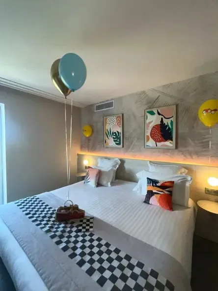 Chambre avec un lit double recouvert d'une couverture à carreaux noirs et blancs, deux tableaux colorés au mur, des ballons et une boîte de chocolats sur le lit