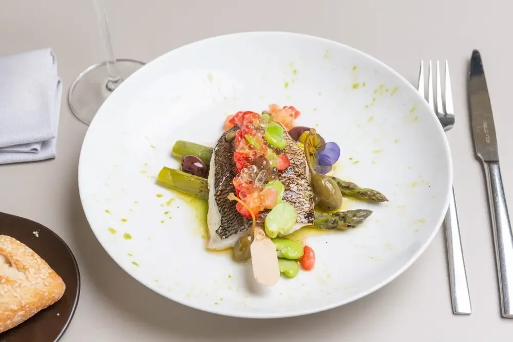 Assiette blanche avec un filet de poisson grillé, des asperges, des légumes variés et une sauce verte, accompagnée d'un couteau, d'une fourchette et d'un verre à vin