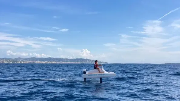 Une personne en maillot de bain se tient sur une plateforme flottante dans la mer, avec un paysage côtier en arrière-plan