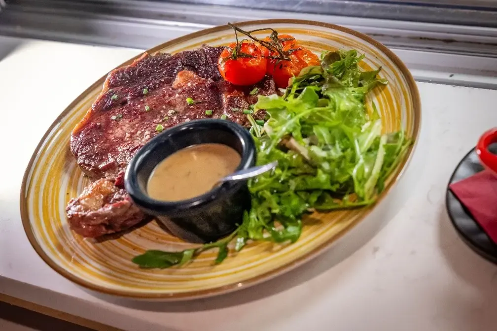 Un plat ovale contient un morceau de viande grillée, une petite salade verte, des tomates cerises et un ramequin de sauce