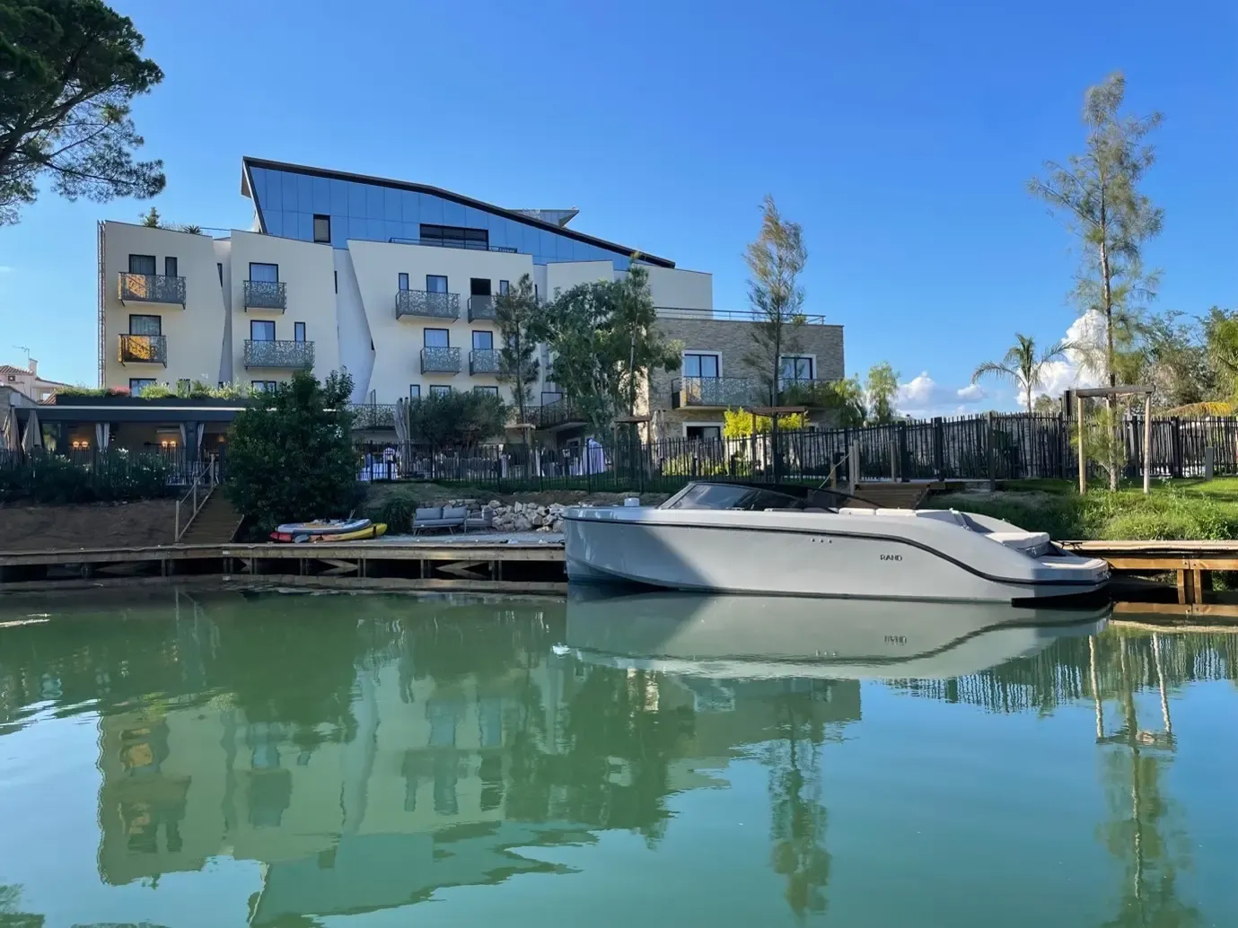 Un bâtiment moderne avec des balcons et des fenêtres vitrées se reflète dans l'eau calme d'un canal, avec un bateau blanc amarré à un quai en bois