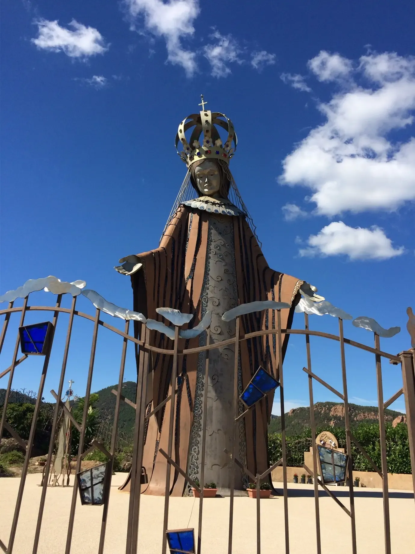 Statue métallique d'une figure religieuse couronnée, entourée d'une structure en fer avec des éléments en verre coloré, sous un ciel bleu avec des nuages