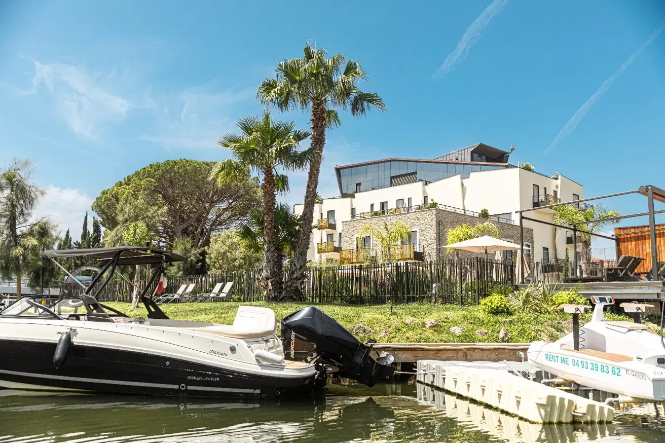 Deux bateaux amarrés devant une maison moderne avec jardin, palmier et terrasse en bord de canal sous un ciel bleu